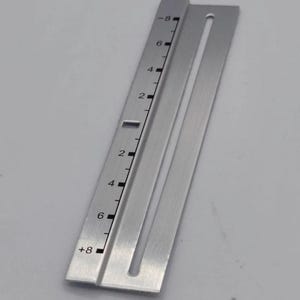 Puede incluir: Una regla de metal plateado con marcas y números negros. La regla tiene una ranura en el centro y medidas de -8 a +8. La herramienta se utiliza probablemente para mediciones precisas.