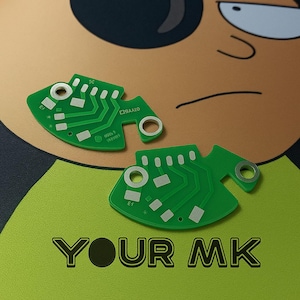 Könnte beinhalten: Zwei grüne Leiterplatten mit silbernen Komponenten und einer einzigartigen fischartigen Form. Die Platinen haben komplizierte silberne Pfade und Text. Der Hintergrund ist ein Cartoon-Bild mit dem Text "YOUR MK" in Schwarz.