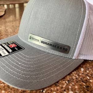 May include: Gray Richardson Trucker hat with a white mesh back. The hat features a metal plate with the text "Allison Transmission DURAMAX 6.6 HD". The hat has a curved brim and a stitched design.