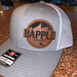 Puede incluir: Gorra de camionero gris con parte trasera de malla blanca y visera curva. La gorra presenta un parche de cuero marrón con el texto "BAPPLE LIGHT" y un gráfico de montaña. La gorra tiene una correa ajustable y una etiqueta.