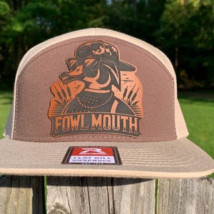Pode incluir: Um boné de beisebol marrom e bege com um remendo de couro marrom apresentando um pato de desenho animado usando óculos de sol e um chapéu. O remendo diz "Fowl Mouth" em letras pretas.