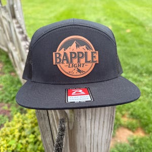 May include: A black snapback cap with a flat bill and a mesh back. The front features a circular tan leather patch with a mountain graphic and the text BAPPLE LIGHT. A rectangular sticker on the bill reads RICHARDSON FLAT BILL SNAPBACK.
