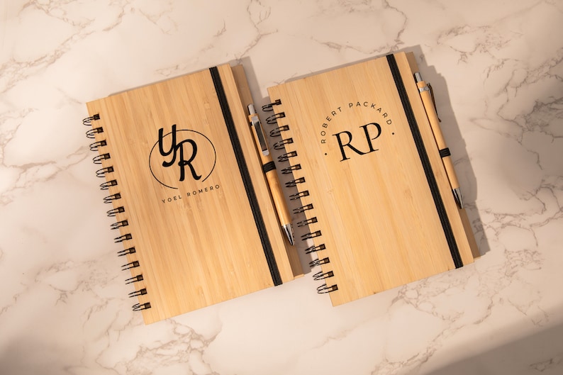 K&ouml;nnte beinhalten: Zwei Holzjournale mit schwarzen Gummib&auml;ndern und passenden Stiften. Das erste Journal hat einen schwarzen Kreis mit den Initialen "YR" und dem Text "Yoel Romero". Das zweite Journal hat die Initialen "RP" und den Text "Robert Packard".