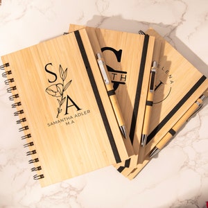 May include: A stack of four wooden journals with black elastic closures and personalized engravings. Each journal has a matching pen with a silver clip. The journals are on a white marble surface.