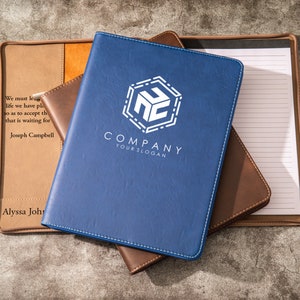 May include: Three leather-bound notebooks, one blue, one brown, and one tan. The blue notebook has a white logo with the text "COMPANY YOURSLOGAN". The tan notebook has the text "We must learn from life we have planned to as to accept the fact that is waiting for us. Joseph Campbell" and "Alyssa John".
