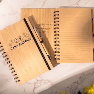 K&ouml;nnte beinhalten: Ein braunes Bambus-Notizbuch mit einem schwarzen Band und einem Stift. Das Notizbuch tr&auml;gt eine personalisierte Inschrift mit dem Text "CS CARA STEWART". Das Notizbuch ist auf einer leeren Seite mit liniertem Papier ge&ouml;ffnet. Der Stift ist ein Bambusstift mit einer silbernen Spitze.