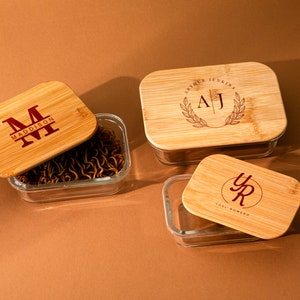 May include: Three glass containers with bamboo lids. Each lid has a personalized engraved design with initials and names. The containers are perfect for storing food or snacks.