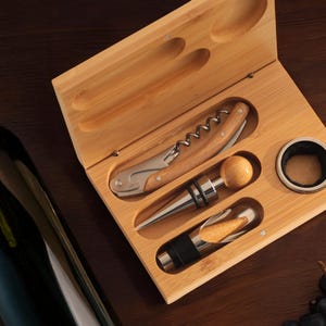 May include: A wooden box with a set of wine tools, including a corkscrew, a bottle stopper, and a foil cutter. The box is open and the tools are visible. There is a bottle of wine and grapes in the background.
