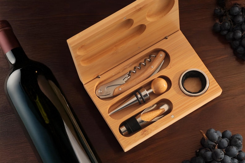 May include: A wooden box with a set of wine tools, including a corkscrew, a bottle stopper, and a foil cutter. The box is open and the tools are visible. There is a bottle of wine and grapes in the background.