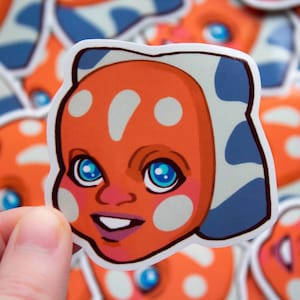 以下が含まれることがあります： オレンジ色の肌、青と白の模様、大きな青い目をした漫画のキャラクターのステッカー。キャラクターは笑顔です。他の同様のステッカーが背景に見えます。