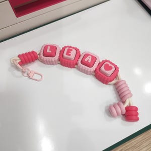 Può includere: Un portachiavi con un cordino bianco infilato con perline rosa e rosa chiaro. Quattro perline quadrate mostrano le lettere L, E, A e un simbolo a forma di cuore. Il portachiavi include un moschettone rosa e perline cilindriche alle estremità.