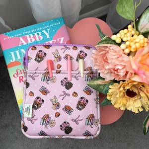 May include: A pink tablet case with a repeating pattern of cartoon dogs, books, and bows. The case has a front pocket with three pens and a purple zipper. A book titled "Abby Jimenez" is visible.