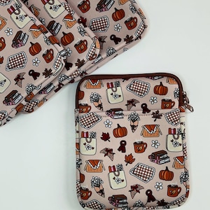 Może przedstawiać: Jasnor&oacute;żowe etui na laptopa i pasujące akcesoria z powtarzającym się wzorem dyni, książek, kwiat&oacute;w i kubk&oacute;w. Etui ma brązowy zamek błyskawiczny i wykończenie. Projekt nawiązuje do jesiennej tematyki.