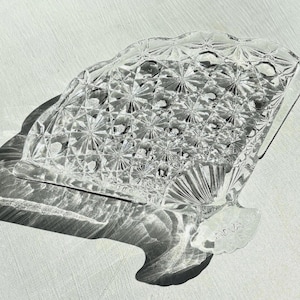 Peut inclure: Un plat en verre taillé transparent avec un motif géométrique, projetant une ombre détaillée sur une surface blanche. Le plat a un bord décoratif festonné et une forme unique, suggérant une pièce vintage.