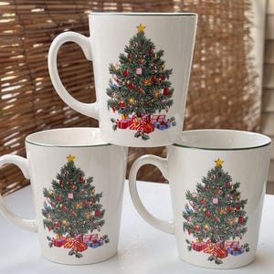 Op de afbeelding: Drie witte keramische mokken met een kerstboomontwerp. Elke mok heeft een groen omrande rand en handvat, met een versierde boom en ingepakte cadeaus. De mokken zijn gestapeld op een wit oppervlak, met een geweven achtergrond.