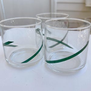 May include: Three clear glass tumblers with a green diagonal stripe design. The glasses have a ribbed texture and are set on a white surface. The glasses are cylindrical and appear to be vintage.