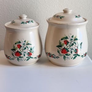May include: Two cream-colored ceramic canisters with matching lids, each featuring a painted pattern of red strawberries, green leaves, and small white flowers. The canisters have a rounded shape and a smooth, glossy finish on the exterior surface.