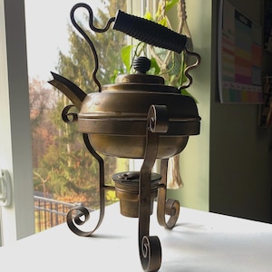 May include: A vintage brass kettle with a black handle and a decorative stand. The kettle is on a white surface.