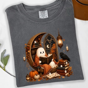 Może przedstawiać: Ciemnoszary t-shirt z grafiką przedstawiającą ducha siedzącego na krześle w kształcie półksiężyca i czytającego książkę. Wzór zawiera czarnego kruka, stosy książek, dynie, wiszącą latarnię, świece i jesienne liście. Koszulka posiada metkę Comfort Colors.