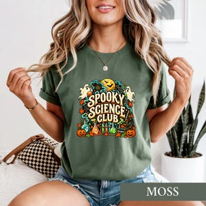 Könnte beinhalten: Ein olivgrünes T-Shirt mit einem farbenfrohen Grafikdruck und dem Text "SPOOKY SCIENCE CLUB". Das Design zeigt Illustrationen von Geistern, Kürbissen, Laborbechern, einem Mikroskop, einem Vollmond und Fledermäusen in Orange-, Blau- und Gelbtönen.