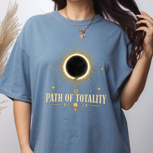 Può includere: Una t-shirt blu a maniche corte con la grafica di un'eclissi solare nera circondata da raggi e stelle dorate. Sotto l'eclissi, il testo PATH OF TOTALITY è scritto in lettere maiuscole dorate, accompagnato da piccole icone delle fasi lunari.
