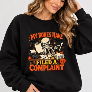 May include: A black crewneck sweatshirt featuring a graphic of a skeleton sitting at a desk with a typewriter, a coffee mug, and papers. The text reads "MY BONES HAVE FILED A COMPLAINT" in orange letters, accented by autumn leaves, bats, and a jack-o'-lantern.