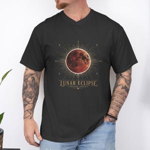 Könnte beinhalten: Ein schwarzes Kurzarm-T-Shirt mit einer Grafik eines roten Blutmondes, der von strahlenden goldenen Linien und Sternen umgeben ist. Unter dem Mond steht der Text LUNAR ECLIPSE und BLOOD MOON in einer dekorativen Serifenschrift mit kleinen Sternen.