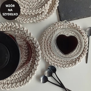 May include: Three beige crocheted coasters with a heart-shaped glass cup filled with black coffee on one of the coasters. The coasters are on a white surface with three black metal spoons.