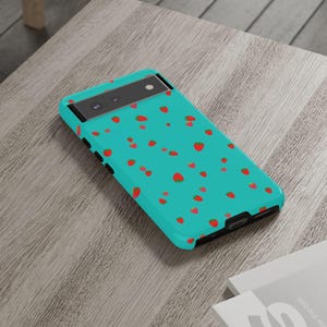 May include: A turquoise phone case with a pattern of red strawberries. The case is on a light brown wooden table. The phone case has a black camera bar and black edges.