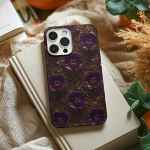 May include: A smartphone case featuring a floral design with purple pansies on a dark brown background. The case is resting on a closed book with a cream-colored cover. The phone case has a camera module with multiple lenses.