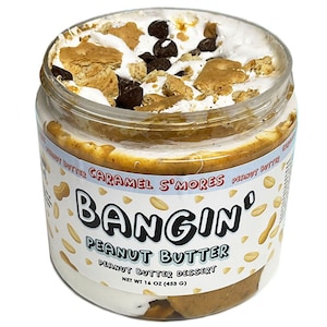 May include: A clear plastic jar of Bangin' Peanut Butter, Caramel S'mores flavour, containing 453 g of peanut butter dessert. The top is filled with white marshmallow cream, dark chocolate chips, and crushed graham cracker pieces.