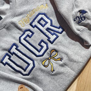 May include: A heather gray sweatshirt with the text "UCR" in blue and white, and "Highlanders" in gold. A small yellow bow and a brown teddy bear patch are also visible. The sleeve features a graduation cap and the number "26".