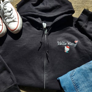 May include: Black zip-up hoodie with a white embroidered Hello Kitty design. The hoodie has a hood and a zipper closure.