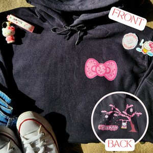 May include: Black hooded sweatshirt with a pink bow with cherry blossoms on the front and a pink Hello Kitty design on a cherry blossom tree on the back.