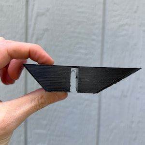 Op de afbeelding: Een zwart, hoekig object met een trapeziumvorm, vastgehouden door een hand. Het object lijkt 3D-geprint te zijn, met zichtbare laaglijnen. Een witte plakstrip is in het midden zichtbaar. Het ontwerp van het object suggereert een mogelijke lade of houder.