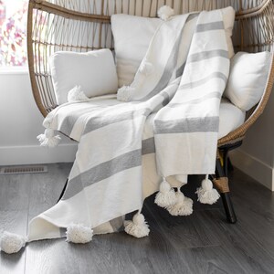 May include: A white and grey striped throw blanket with white pom-poms on a wicker egg chair. The chair is in a room with a wooden floor.