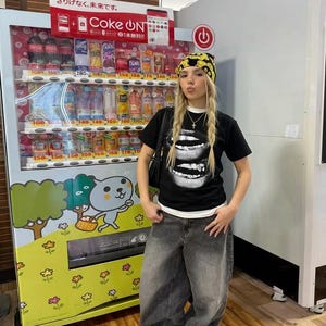 May include: A person wearing a black t-shirt with a white lip graphic, paired with oversized gray jeans and a yellow patterned beanie. A vending machine with Japanese text and various beverages is in the background.