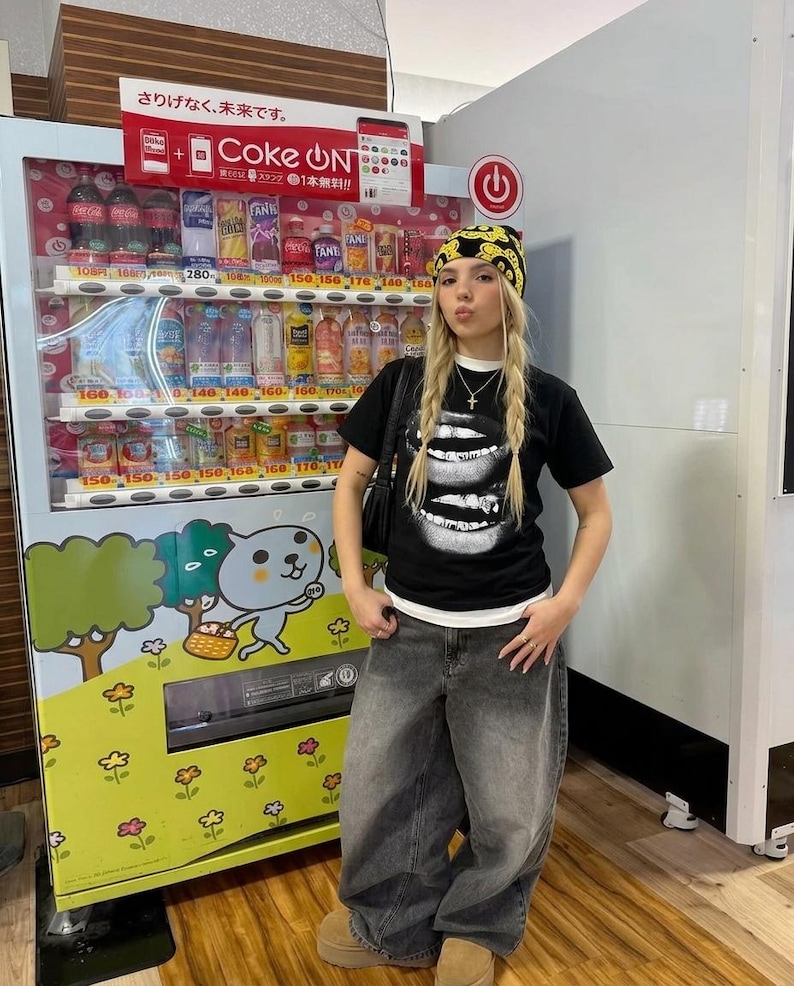 May include: A person wearing a black t-shirt with a white lip graphic, paired with oversized gray jeans and a yellow patterned beanie. A vending machine with Japanese text and various beverages is in the background.