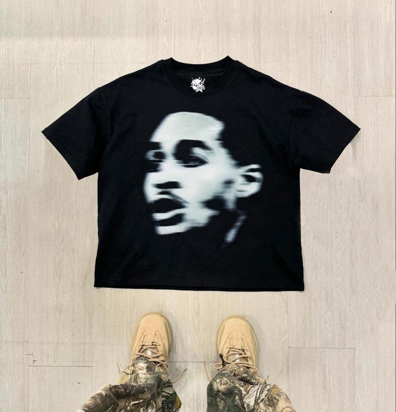 Op de afbeelding: Zwart T-shirt met een witte grafische print van een gezicht. Het gezicht is wazig en de afbeelding is in zwart-wit.