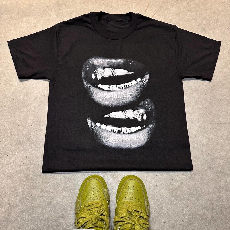 May include: Black t-shirt featuring a grayscale image of two sets of smiling lips with prominent teeth. The shirt is paired with olive green sneakers. The design is centered on the shirt, creating a bold visual statement.