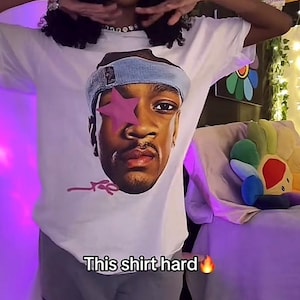 Puede incluir: Camiseta blanca con un retrato gr&aacute;fico de una persona con una estrella rosa sobre un ojo y una diadema azul. El texto "This shirt hard" est&aacute; impreso en la camiseta. La persona lleva un pantal&oacute;n de ch&aacute;ndal gris.