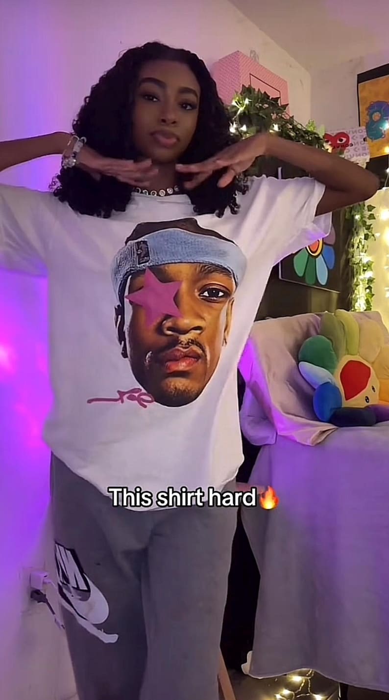 Puede incluir: Camiseta blanca con un retrato gr&aacute;fico de una persona con una estrella rosa sobre un ojo y una diadema azul. El texto "This shirt hard" est&aacute; impreso en la camiseta. La persona lleva un pantal&oacute;n de ch&aacute;ndal gris.