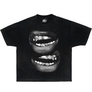 May include: A black short-sleeve t-shirt featuring a graphic print of two sets of lips stacked vertically. The lips are rendered in grayscale and show teeth adorned with metallic, diamond-like grillz that catch the light.