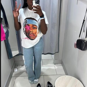 Puede incluir: Camiseta blanca con un gr&aacute;fico de una persona con una diadema azul. La persona lleva vaqueros azul claro y zapatillas deportivas blancas y negras. La imagen est&aacute; tomada en un probador.