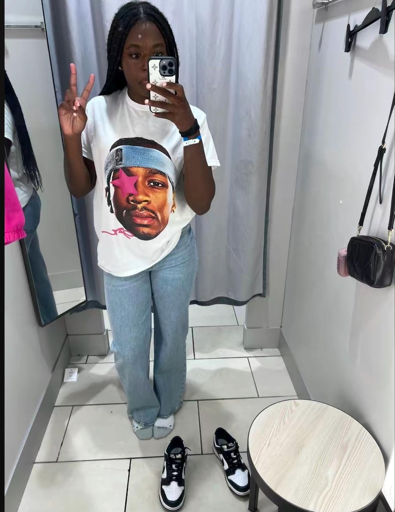 Puede incluir: Camiseta blanca con un gr&aacute;fico de una persona con una diadema azul. La persona lleva vaqueros azul claro y zapatillas deportivas blancas y negras. La imagen est&aacute; tomada en un probador.