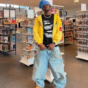 May include: A person wearing a blue beanie, yellow zip-up hoodie, black graphic t-shirt, light blue baggy jeans, and tan work boots. The t-shirt features a graphic of white lips. A gold chain hangs from the jeans.