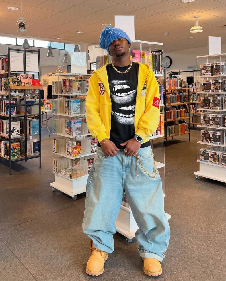 May include: A person wearing a blue beanie, yellow zip-up hoodie, black graphic t-shirt, light blue baggy jeans, and tan work boots. The t-shirt features a graphic of white lips. A gold chain hangs from the jeans.