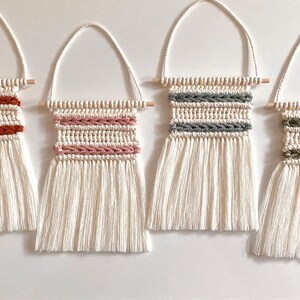Peut inclure: Quatre petites tentures murales en macramé avec des chevilles en bois et des franges blanches. Les tentures présentent des rayures tissées de différentes couleurs : rouge, rose, gris et vert.