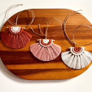 May include: Three handmade macrame wall hangings with fringe, each featuring a different colour combination: rust, pink, and white. The hangings are displayed on a wooden surface.