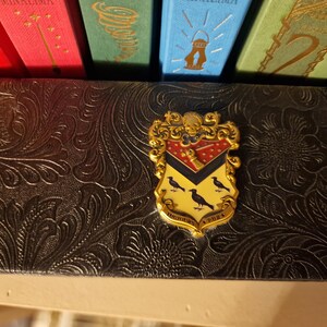 May include: A gold-colored crest pin with a red, black, and yellow shield design, featuring three black birds. The pin is set against a dark, textured background, with colorful book spines visible above.